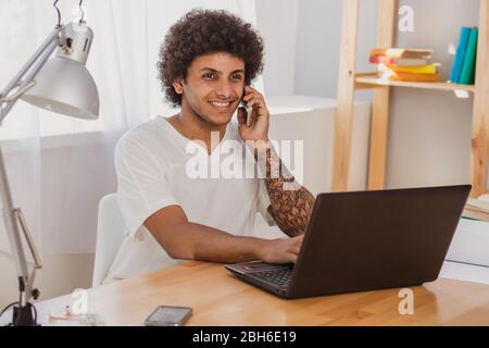 Curly h Curly haired man working on laptop and talking on the mobile phone while sitting at his working place at home Stock Photo