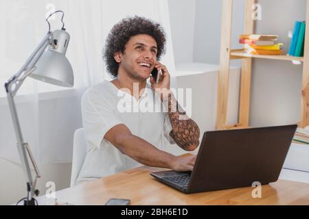 Cheerful african man working on laptop and talking on the mobile phone while sitting at his working place in modern office Stock Photo