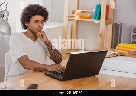 Pensive man, with curly hair and dressed informally,intently looking at laptop while sitting at his working place at home Stock Photo