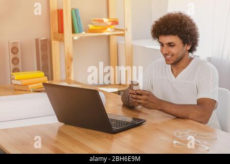 handsome young man of arabian ethnicity, smiling and looking to his phone, chatting, sitting at his workplace with laptop . Stock Photo