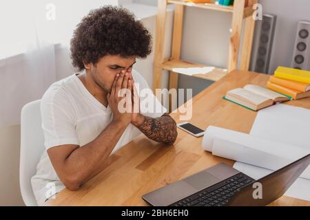 Curly haired stylish man sick and tired after work , sitting at his workplace, massaging his nose . Stock Photo