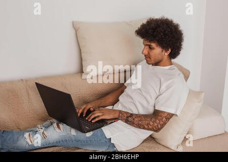 Young man, a mulatto with curly hair, relaxed on the couch using a laptop, at home Stock Photo