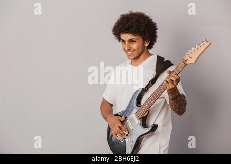Positive curly haired man, expressive plays the bass guitar,on gray background. Cheerful musician. Stock Photo