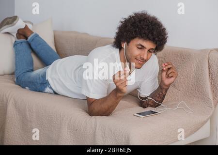 Curly haired stylish man eyes closed lying on the couch at home, enjoy the music. Stock Photo