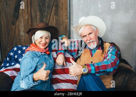 Elderly couple dressed in cowboy hats, showing thumbs up, smiling and looking at camera. Behind, on the couch lies the American flag, the celebration Stock Photo