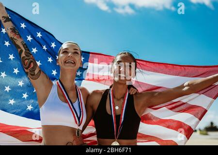 Two woman athletes holding up american flag outdoors. Female USA runners celebrating victory on running track in stadium. Stock Photo