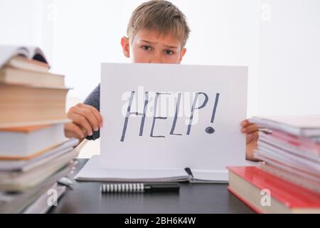 Sad tired frustrated boy sitting at the table with many books and holding paper with word Help. Learning difficulties, education concept. Stock Photo