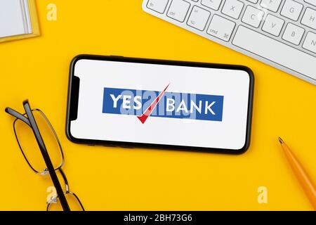 A smartphone showing the Yes Bank logo rests on a yellow background along with a keyboard, glasses, pen and book (Editorial use only). Stock Photo