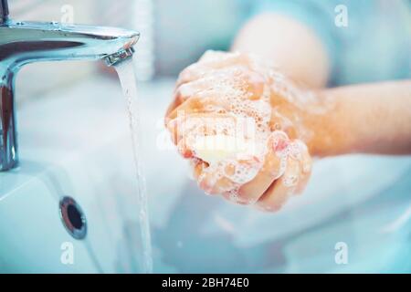 A man carefully washes his hands with soap and thick foam in the bathroom over the sink, from the tap of which water flows.