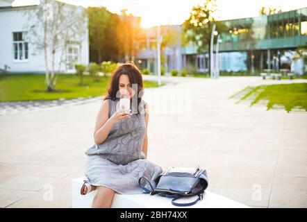 Happy pregnant woman holding cell phone while sitting on park bench outdoors Stock Photo