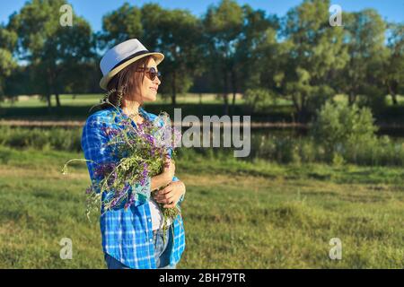 Country portrait of an adult beautiful woman in hat with bouquet of wildflowers. Green natural landscape, spring summer meadows, female looks away, co Stock Photo