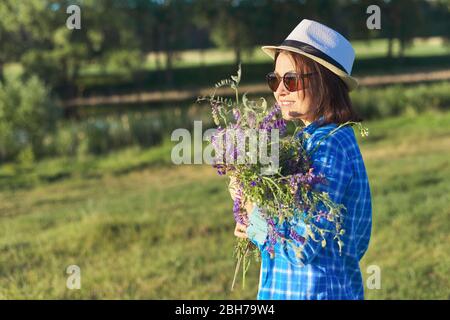 Country portrait of an adult beautiful woman in hat with bouquet of wildflowers. Green natural landscape, spring summer meadows, female looks away, co Stock Photo