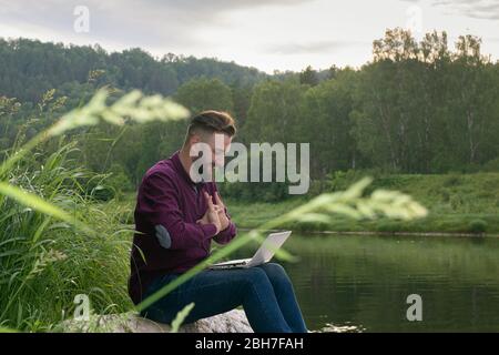 Young man working with laptop sitting by river on summer day. Programmer surrounded by beautiful nature getting ready for work, stretching fingers. Fr Stock Photo