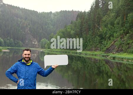 Young smiling man with blank sign board in hands against beautiful nature background. Tourism, outdoors recreation, active style of life Stock Photo