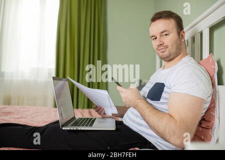 Adult man lying in his bed while he is working from home on his laptop looking on the camera. Papers or bills in his hand and a phone Stock Photo