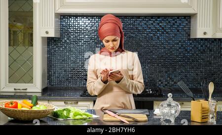 beautiful Muslim woman in hijab surfs Internet using modern smartphone and looking for salad recipe in kitchen closeup Stock Photo