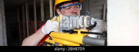 Builder in helmet holds heavy tool, drills wall Stock Photo