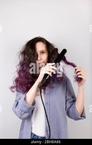 Young girl straightens curly hair of Flat Iron. Coloring purple hair. Gray background Stock Photo