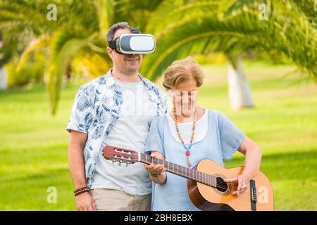 nice happy couple in rare situation - mother and son aged with guitar and goggles headset - modern and ancient way to entertain people - playful and crazy leisure activity - Stock Photo
