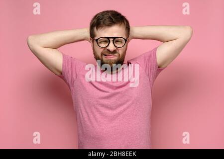 Bearded man with glasses is not in the spirit, he grimaces in exasperation, putting hands behind head, on a pink background. Stock Photo