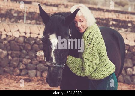 Vintage colors style for atractive blonde woman hugging a black quiet horse in outdoor leisure activity - animals best friend odd forever - romantic concept for young female Stock Photo