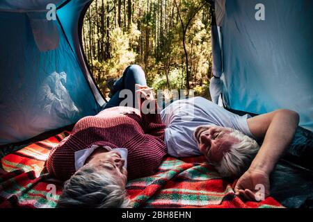 adult senior couple rest lay down inside a tent in free wild camping in the forest for alternative travel and lifestyle. Love forever together concept for man and woman taking hands andd look eachother - outdoor nature activity Stock Photo