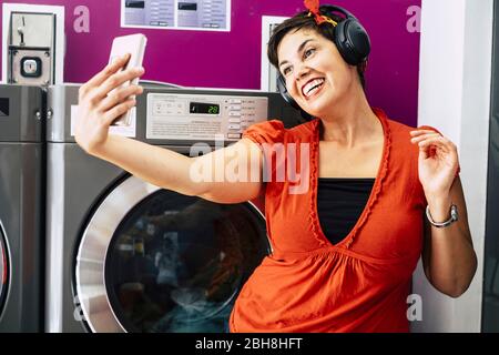 Cheerful nice brunette taking selfie with smart phne at the laundromat while listen music and wait for her dress washer - young millennial people living in the city concept - laundry activity business Stock Photo
