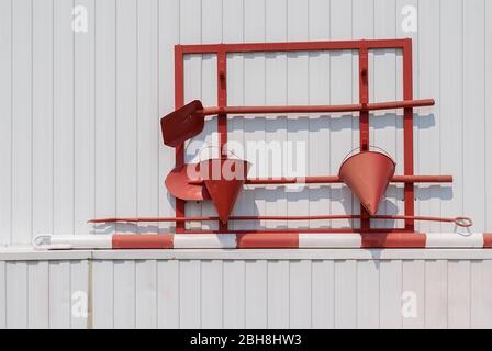 a red fire shield on a white metal wall with buckets, shovels, and primitive, simple fire tools Stock Photo