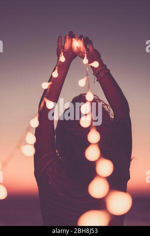 Motivational inspirational concept image with girl from back holding yellow bulb lights to the sky - pink coloured style filter - hope and feeling - lifestyle for happy people - sunset in background Stock Photo