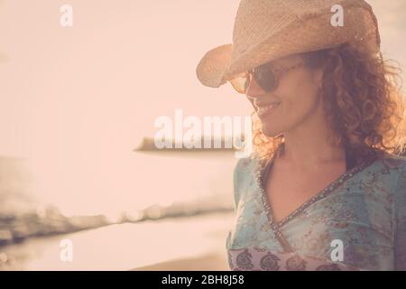 beautiful and wonderful blonde curly hair model middle age with cowboy hat smile and pose at the beach with golden sunset in the background. colored and freedom independence strong woman concept Stock Photo