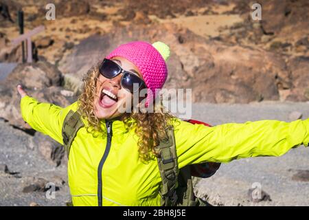 Young beautiful woman laugh and smile with joyful and open arms enjoying the alternative hiking vacation in outdoor mountains in summer - sunny day and funny pink hat for the cold temperature