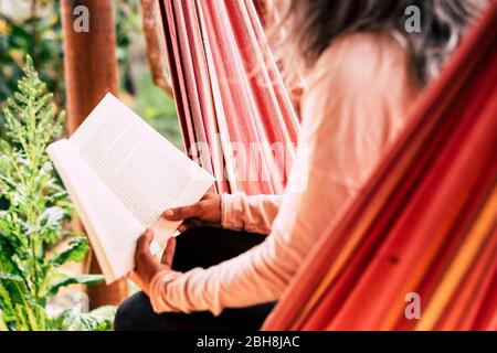 Reading a paper book old style concept to relax and study - aged caucsian woman with white hair sitting on an hammock outdoor enjoy the leisure activity Stock Photo