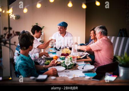 Dinner time in friendship with different ages people all together having fun and enjoying the night with smiles and happiness. Mobile phones defocused in for technology modern concept Stock Photo