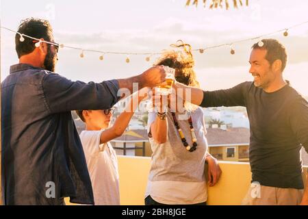 Group of friends toasting and clinking glasses with beer and orange juice together during the sunset on the roof terrace with city view - mixed ages and generations having fun celebrating