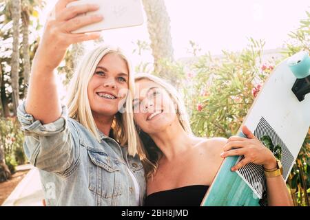 Happy coule of young caucasian attractive girls taking selfie picture with modern phone - braket for perfect teeth and youthful concept - people in summer outdoor leisure activity together Stock Photo