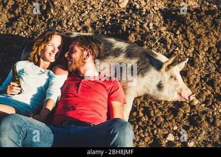 Diversity animal love pet therapy concept with young beautiful couple of young peope lay down on a nice cheerful pig sleeping on the ground in a sunny day - alternative lifestyle with nature rural Stock Photo
