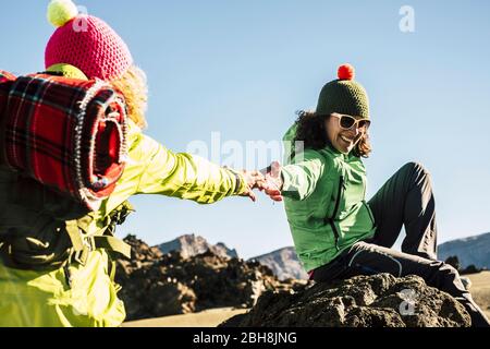 Couple of happy woman helping eachother to arrive to the top of the mountain - enjoy the outdoor trekking hiking activity - clear blue sky in background and concept of team and work together in friendship Stock Photo