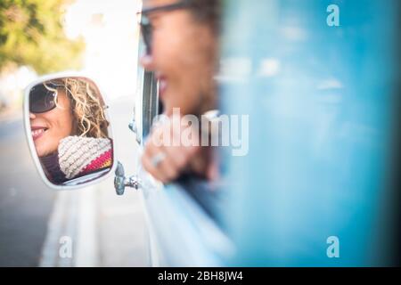 Cheerful lady smile and travel on a old blue vehicle and looking herself on the back mirror - summer colors and roadtrip travelimg concept - attractive curly lady enjoying drive vehicle Stock Photo