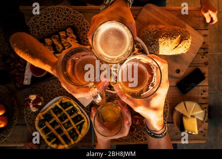 Top view of hands with beers cheering and having fun together - friends at dinner celebrating - wood table with a lot of food viewed from above - friendship and event party concept Stock Photo