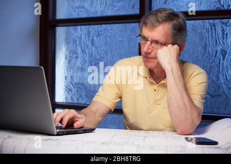 Senior pensive businessman or engineer working from home at a desk with laptop, plan and mobile phone - focus on the face Stock Photo