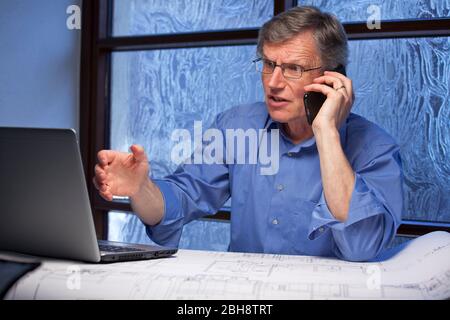 Mature angry businessman talking on mobile phone and looking at the monitor of a laptop - focus on the face Stock Photo