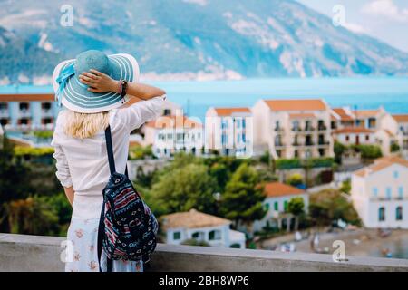 Tourist woman admiring view of colorful tranquil village Assos on morning. Young stylish female model wearing blue sunhat and white clothes enjoying summer travel vacation in Kefalonia, Greece. Stock Photo