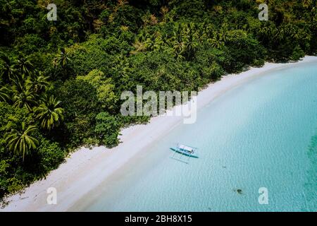 Aerial drone view of a beautiful secluded deserted tropical beach. Lonely boat in turquoise lagoon in front of rainforest jungle. Cadlao Island, El Nido, Palawan.