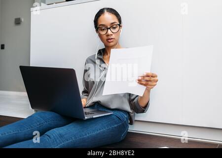 Young woman sitting on floor at kitchen counter with document in hand and using laptop Stock Photo