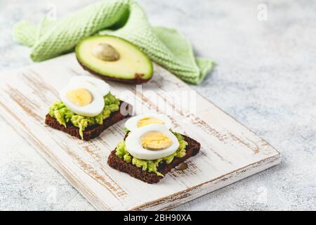 Healthy toasts on rye bread with avocado and boiled eggs on white wooden cutting board. Delicious appetizer, ideal as an aperitif. Delicious breakfast Stock Photo