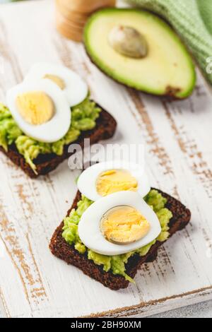 Healthy toasts on rye bread with avocado and boiled eggs on white wooden cutting board. Delicious appetizer, ideal as an aperitif. Delicious breakfast Stock Photo