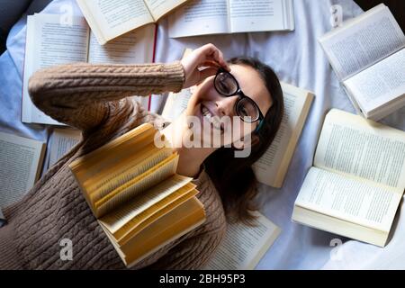 Woman with glasses lying on the bed next to many books. Stock Photo