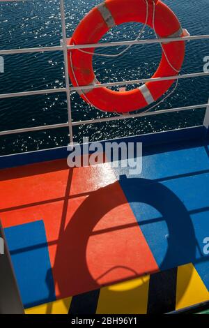 orange lifebuoy hangs on railing of a ferry, shadows on colorful ground, strong color contrast, red and blue Stock Photo