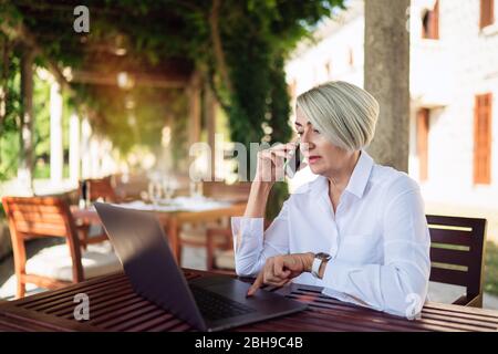 Mature woman using mobile phone while resting at the cafe with laptop computer Stock Photo