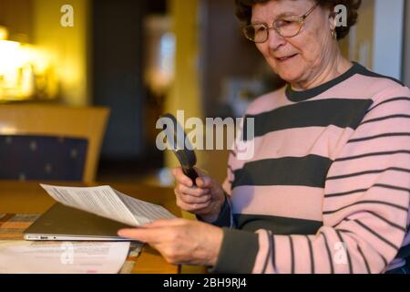 Happy senior woman smiling while reading paper with magnifying glass in the dining room Stock Photo